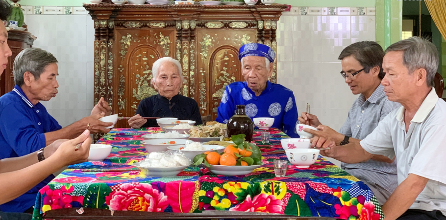 Cảnh mọi người cùng nhau ngồi quanh bàn cỗ, cười nói rộn ràng, thể hiện sự đoàn kết và hạnh phúc.