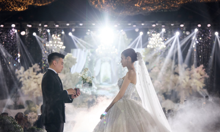 Một lễ cưới sang trọng, cô dâu chú rể nắm tay nhau giữa khung cảnh lung linh ánh đèn.