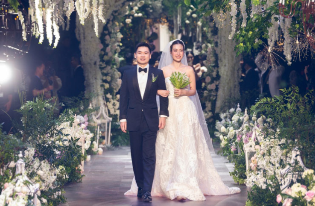 Bức ảnh cặp vợ chồng nắm tay nhau, phía xa là khung cảnh tiệc cưới lung linh.