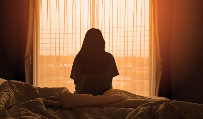 Một người đang ngồi trầm tư cạnh giường bệnh, ánh sáng chiếu nhẹ qua cửa sổ thể hiện sự lo lắng và yêu thương.